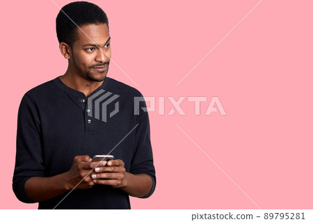Image of handsome guy with dark skin, in black shirt, stands and holds phone in hand, looks aside with uncomprehending, hears unpleasent conversation near him while typing message for his firlfriend. Image of handsome guy with dark skin, in black shirt, stands and holds phone in hand, looks aside with uncomprehending, hears unpleasent conversation near him while typing message for his firlfriend. 89795281
