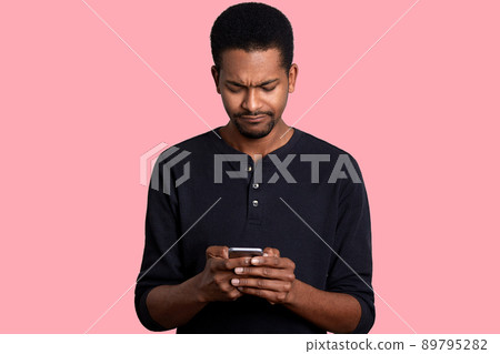 Portrait of attractive African American man with telephone in hands, surfes Internet. Handsome black male looks weather forecast for weekend with sad fecial expression. Modern technology concept. Portrait of attractive African American man with telephone in hands, surfes Internet. Handsome black male looks weather forecast for weekend with sad fecial expression. Modern technology concept. 89795282