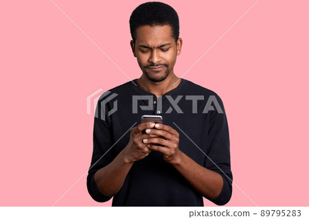 Attractive black man holds mobile phone and looks worried. Black guy reads message with unpleasent news from friend and does not know how to help. Male wears casual jumper, stands against pink wall. Attractive black man holds mobile phone and looks worried. Black guy reads message with unpleasent news from friend and does not know how to help. Male wears casual jumper, stands against pink wall. 89795283