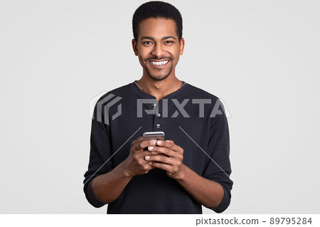 Photo of cheerful dark skinned man with toothy smile, holds modern smart phone, types text message to girlfriend, dressed in casual clothes, isolated over white background. Technology concept Photo of cheerful dark skinned man with toothy smile, holds modern smart phone, types text message to girlfriend, dressed in casual clothes, isolated over white background. Technology concept 89795284