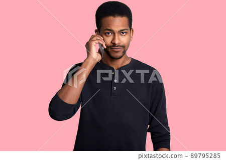 Black man talks on mobile phone, discuses important topic. Handsome African guy with sad expression, looks at camera, poses in photo studio with his smart phone isolated over rose background. Black man talks on mobile phone, discuses important topic. Handsome African guy with sad expression, looks at camera, poses in photo studio with his smart phone isolated over rose background. 89795285