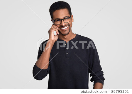 Positive dark skinned Afro American man with cheerful expression, has telephone conversation, discusses something pleasant with friend, organize party together, isolated over white background. Positive dark skinned Afro American man with cheerful expression, has telephone conversation, discusses something pleasant with friend, organize party together, isolated over white background. 89795301
