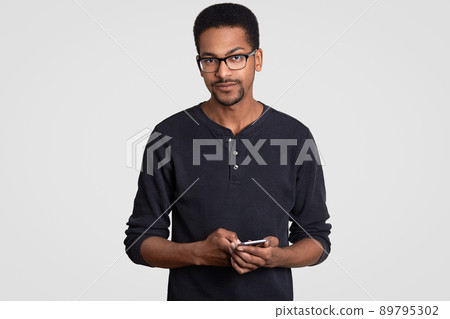 Indoor shot of serious man with dark skin, has confident facial expression, holds modern cellular, texts sms, reads feedback, wears glasses, isolated over white background. Ethnicity, technology Indoor shot of serious man with dark skin, has confident facial expression, holds modern cellular, texts sms, reads feedback, wears glasses, isolated over white background. Ethnicity, technology 89795302