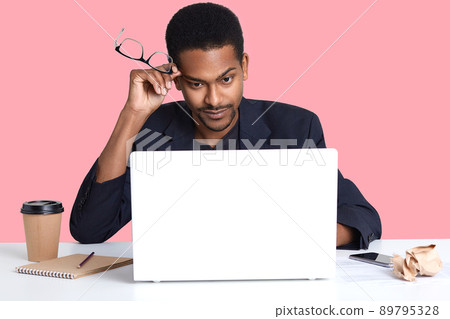 Photo of tired man holds glasses in hands near temple, wears black suit, prepares business project, uses modern portable laptop, isolated over pink wall, erans money in office. People concept. 89795328