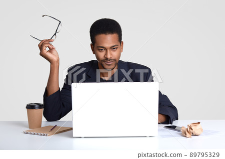 Photo of serious dark skinned man works freelance, keeps eyewear in hands, reads news on internet website, focused into monitor of laptop, wears formal suit, sits in coworking space over white wall Photo of serious dark skinned man works freelance, keeps eyewear in hands, reads news on internet website, focused into monitor of laptop, wears formal suit, sits in coworking space over white wall 89795329