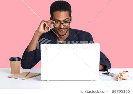 Portrait of dark skinned man works with laptop. Handsome black male sits at desk wears suit, touches his spectacles with hand, looks concentrated at monitor. Online working and technology concept Portrait of dark skinned man works with laptop. Handsome black male sits at desk wears suit, touches his spectacles with hand, looks concentrated at monitor. Online working and technology concept 89795330