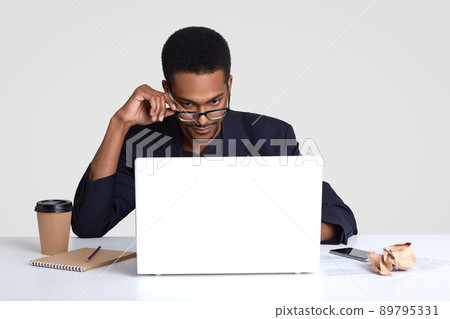 Indoor shot of dark skinned man looks attentively through spectacles, concentrated on graphic, works on new startup project, makes notes in diary, drinks takeaway coffee isolated over white background Indoor shot of dark skinned man looks attentively through spectacles, concentrated on graphic, works on new startup project, makes notes in diary, drinks takeaway coffee isolated over white background 89795331