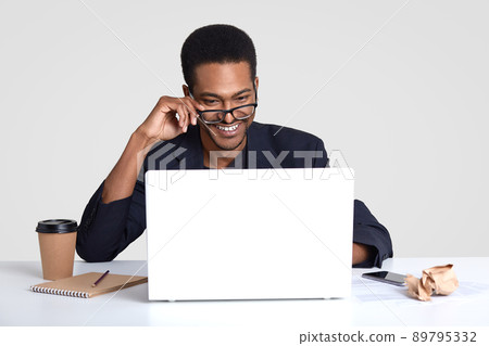 Smiling positive dark skinned man wears transparent glasses, focused into laptop computer, makes distant job, writes down something in notepad, drinks takeout coffee, isolated on white wall. Smiling positive dark skinned man wears transparent glasses, focused into laptop computer, makes distant job, writes down something in notepad, drinks takeout coffee, isolated on white wall. 89795332
