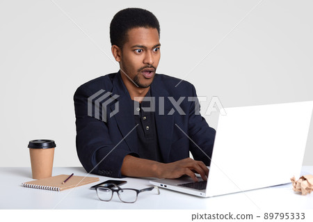 Shocked businessman with black skin, keyboards information on laptop computer, stupefied to fail make presentation, drinks coffee, writes down information in notepad, isolated over white background Shocked businessman with black skin, keyboards information on laptop computer, stupefied to fail make presentation, drinks coffee, writes down information in notepad, isolated over white background 89795333