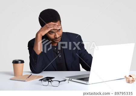 Tired fatigue dark skinned unshaven man feels overworked after long time working, keeps hand on forehead, feels migraine, wears formal suit, uses notepad for writing records, drinks takeaway coffee 89795335