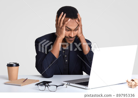 Overworked tired stressful male keeps hands on head, lools with puzzlement, doesnt know how to prepare financial report, wears formal clothes, focused into portable laptop computer, writes notes Overworked tired stressful male keeps hands on head, lools with puzzlement, doesnt know how to prepare financial report, wears formal clothes, focused into portable laptop computer, writes notes 89795336