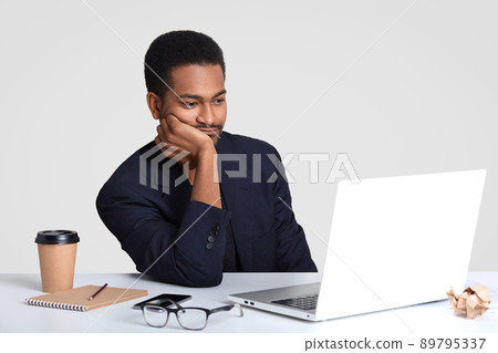 Photo of dark skinned Afro American man keeps hand on cheek, watches attentively tutorial video, prepares for business meeting, has notepad and pen on white desk, wears formal suit, poses indoor Photo of dark skinned Afro American man keeps hand on cheek, watches attentively tutorial video, prepares for business meeting, has notepad and pen on white desk, wears formal suit, poses indoor 89795337