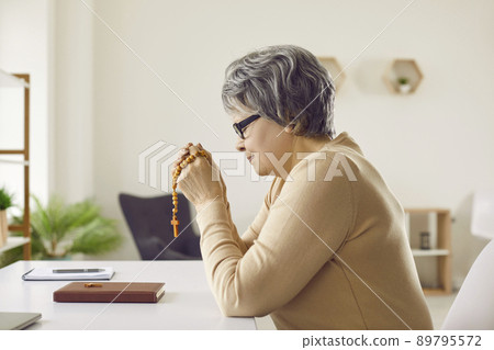Senior woman who belongs to Catholic Christian Church sitting at desk and praying 89795572