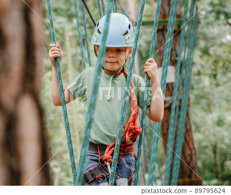 Brave little boy having fun at adventure park and smiling to camera wearing helmet. Scout practicing rappelling. Child - a boy and a girl in the rope park pass obstacles. Brave little boy having fun at adventure park and smiling to camera wearing helmet. Scout practicing rappelling. Child - a boy and a girl in the rope park pass obstacles. 89795624