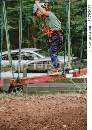Brave little boy having fun at adventure park and smiling to camera wearing helmet. Scout practicing rappelling. Child - a boy and a girl in the rope park pass obstacles. 89795625