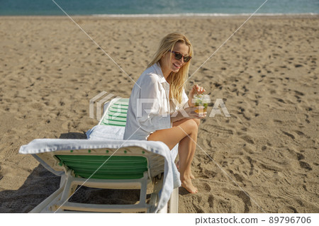Tanned young woman in bikini and shirt with refreshing drink sitting on a deck chair, sunbathing on a beach 89796706