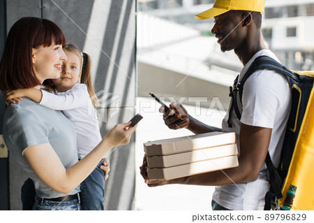 Young smiling woman with a girl at the door, pay by card for online order. Young smiling woman with a girl at the door, pay by card for online order. 89796829