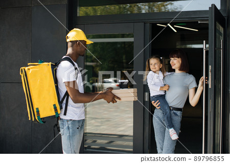Smiling African American delivery man in yellow uniform cap delivering cardboard boxes with pizza Smiling African American delivery man in yellow uniform cap delivering cardboard boxes with pizza 89796855