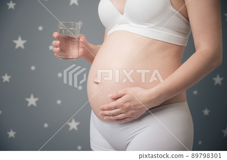 Pregnant woman holding glass with drinking water. Unrecognized pregnant female tenderly touching belly at home. Healthy maternity concept. 89798301