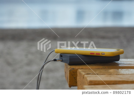 Power Bank charges a smartphone on a wooden bench on the beach with a lake in the background. Power Bank charges a smartphone on a wooden bench on the beach with a lake in the background. 89802178