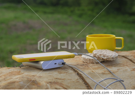The smartphone is charging in nature. A portable charger power bank charges the phone on a tree against the background of a cup of coffee and cookies. The smartphone is charging in nature. A portable charger power bank charges the phone on a tree against the background of a cup of coffee and cookies. 89802229
