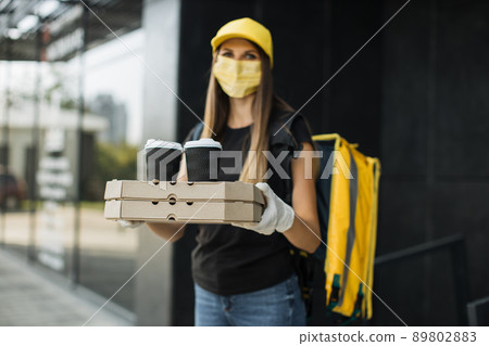 Portrait of delivery woman with yellow cap in face mask and thermal backpack looking at camera Portrait of delivery woman with yellow cap in face mask and thermal backpack looking at camera 89802883