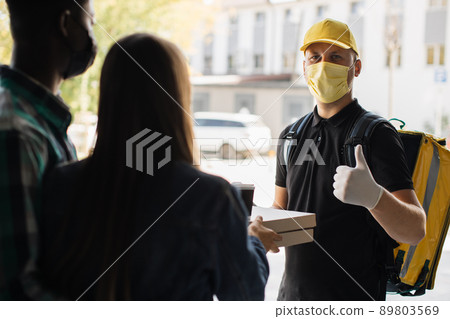 Smilling delivery man in yellow face mask, uniform cap delivering cardboard boxes with pizza 89803569