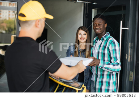 Delivery man in yellow uniform delivering parcel box to recipient - young woman and her husband Delivery man in yellow uniform delivering parcel box to recipient - young woman and her husband 89803618