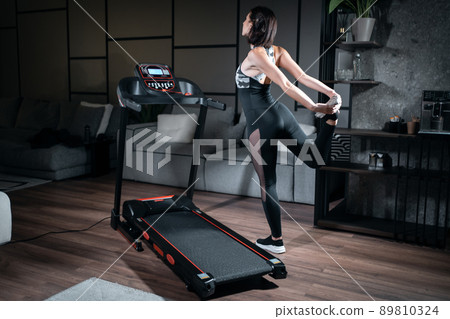 cheerful female stretching and preparing for cardio workout on treadmill at home 89810324