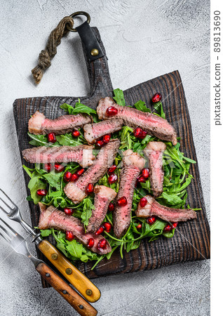 Sliced grilled beef steak with arugula leaves salad on rustic cutting board. White background. Top view Sliced grilled beef steak with arugula leaves salad on rustic cutting board. White background. Top view 89813690
