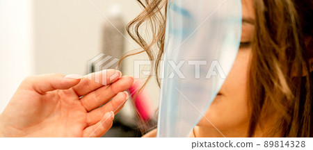 Professional hairdresser styling and fixing with hairspray long woman curly hair covering the face with a protective shield in a beauty salon 89814328