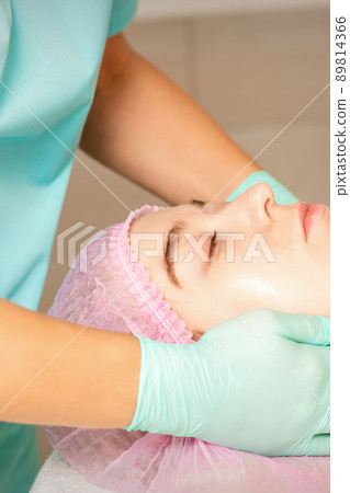 Cosmetologist with gloved hands applies a moisturizing mask with peeling cream on the female face. Facial cosmetology treatment. Procedures for facial care Cosmetologist with gloved hands applies a moisturizing mask with peeling cream on the female face. Facial cosmetology treatment. Procedures for facial care 89814366