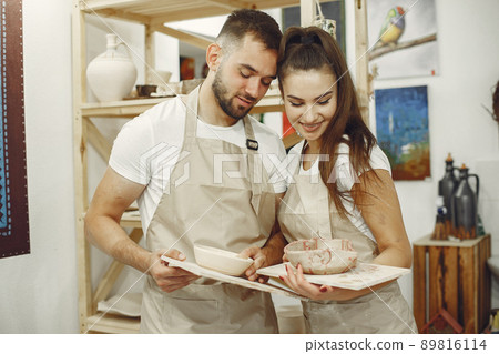 Mutual creative work. Young beautiful couple in casual clothes and aprons. People holds ceramic dishes. 89816114