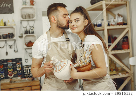 Mutual creative work. Young beautiful couple in casual clothes and aprons. People holds ceramic dishes. 89816152