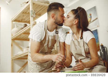 Mutual creative work. Young beautiful couple in casual clothes and aprons. People creating a bowl on a pottery wheel in a clay studio. Mutual creative work. Young beautiful couple in casual clothes and aprons. People creating a bowl on a pottery wheel in a clay studio. 89816420