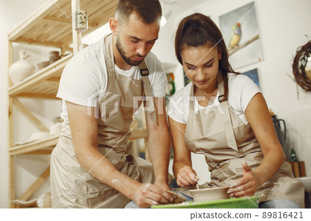 Mutual creative work. Young beautiful couple in casual clothes and aprons. People creating a bowl on a pottery wheel in a clay studio. Mutual creative work. Young beautiful couple in casual clothes and aprons. People creating a bowl on a pottery wheel in a clay studio. 89816421