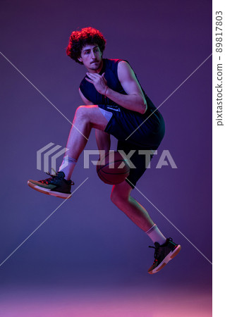 Dynamic portrait of professional basketball player jumping with ball isolated on purple background in neon. Concept of sport, art, creation, competition. Dynamic portrait of professional basketball player jumping with ball isolated on purple background in neon. Concept of sport, art, creation, competition. 89817803