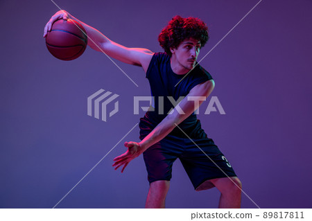 Dynamic portrait of professional basketball player jumping with ball isolated on purple background in neon. Concept of sport, art, creation, competition. Dynamic portrait of professional basketball player jumping with ball isolated on purple background in neon. Concept of sport, art, creation, competition. 89817811