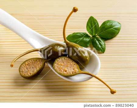 Pickled caper berries in a white spoon and basil leaf on a wooden background. 89823040