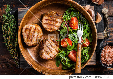 Roasted pork medallions steaks from tenderloin fillet with vegetable salad in wooden plate. Wooden background. Top view Roasted pork medallions steaks from tenderloin fillet with vegetable salad in wooden plate. Wooden background. Top view 89823576