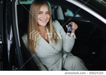 Portrair of attactive young bussines woman sitting in car in car showroom. Woman choosing new car. Beautiful blond hair female in suit and show car keys 89824183