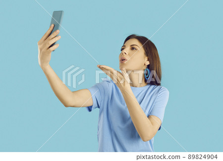 Beautiful young woman holding her mobile phone, blowing an air kiss and taking a selfie 89824904