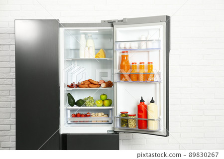 Open refrigerator filled with food near white brick wall 89830267