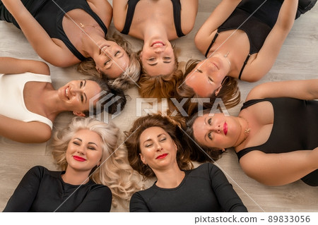 Seven happy women lie on their backs on the floor with their heads pressed together. 89833056