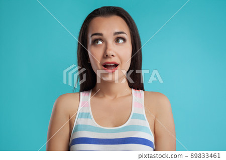 Brunette woman with long hair, dressed in colorful striped shirt, posing against blue studio background. Sincere emotions. Close-up. 89833461