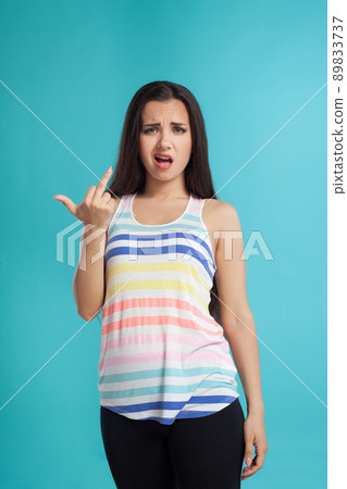 Brunette woman with long hair, dressed in striped shirt and black leggings, posing against blue studio background. Sincere emotions. Close-up. 89833737
