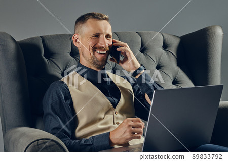 Man with stylish mustache, dressed in black shirt and beige vest is sitting on dark sofa, using his laptop and smartphone. Grey background, close-up. 89833792