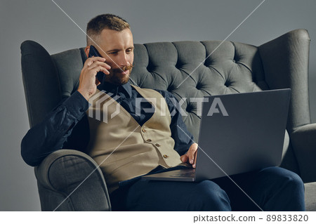 Man with stylish mustache, dressed in black shirt and beige vest is sitting on dark sofa, using his laptop and smartphone. Grey background, close-up. Man with stylish mustache, dressed in black shirt and beige vest is sitting on dark sofa, using his laptop and smartphone. Grey background, close-up. 89833820
