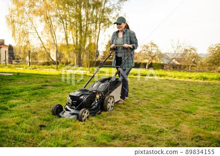 Pushing grass trimming lawnmower. caucasian 30s woman using electric lawn mower while working at garden. 89834155