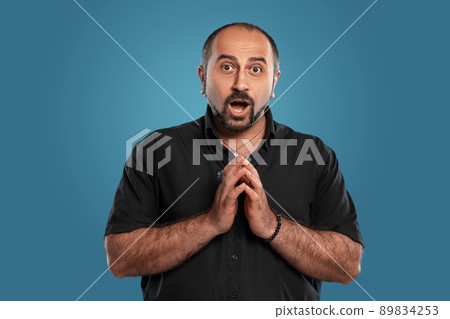 Close-up portrait of a brunet middle-aged man with beard, dressed in a black t-shirt and posing against a blue background. Close-up portrait of a brunet middle-aged man with beard, dressed in a black t-shirt and posing against a blue background. 89834253